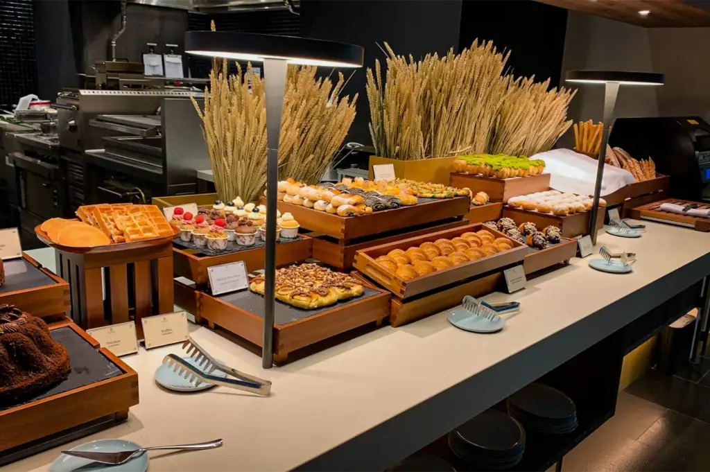 A high-angle, medium shot captures an elegant breakfast pastry buffet spread inside a restaurant kitchen or hotel dining room, representing the file "4-Inside Pan Pacific Orchard Luxury.webp". Arranged neatly on wooden blocks and risers across a long white counter, the buffet features an assortment of baked goods including fresh croissants, frosted cupcakes, danishes, pastries with green icing, sliced cakes, waffles, and pancakes. Tall decorative bunches of dried wheat stalks sit behind the pastries against a dark kitchen background, while two modern, minimalist black table lamps cast warm, focused light over the selection. Small white labeled placards are placed in front of each dish alongside metal serving tongs and blue ceramic plates, creating a clean and sophisticated presentation.